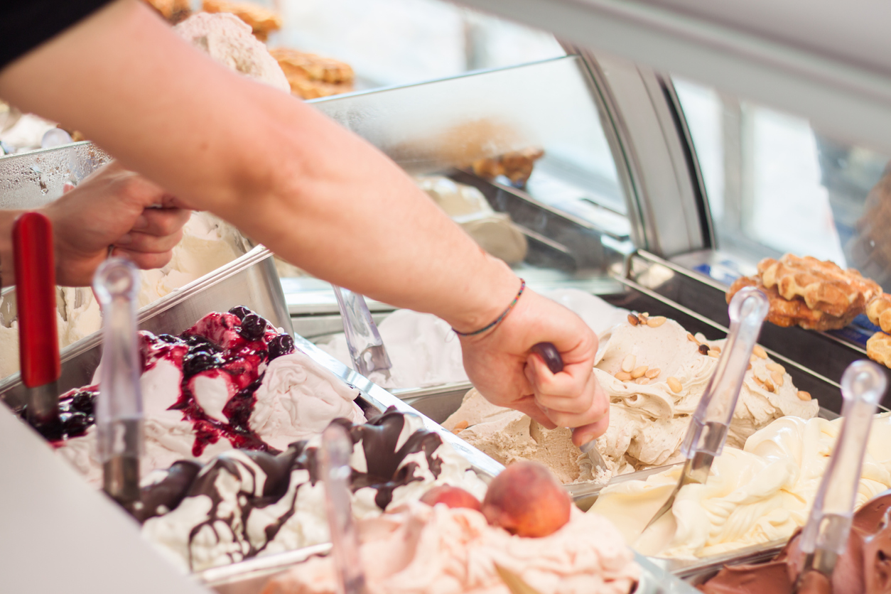 This is a photo of a man who sell ice-cream in ice-cream shop Italian gelateria. The man is filling waffle with an ice cream with his hand. The display case inside with ice creams is seen in the background.