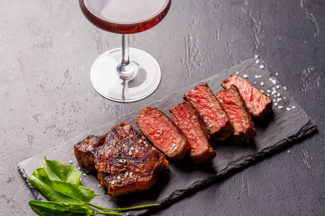A perfectly cooked sliced striploin steak with a juicy, tender texture paired with red wine on a dark background. A gourmet meal for steak and wine lovers