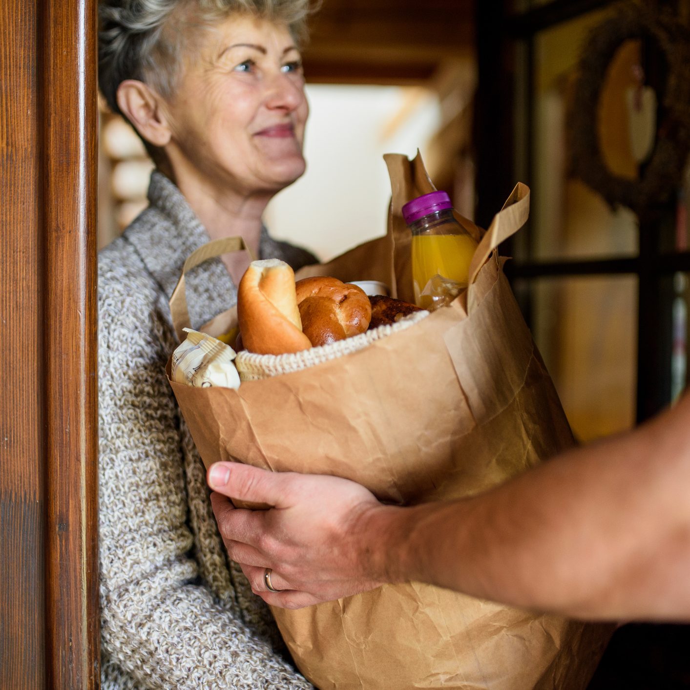 Unrecognizable courier delivering shopping in paper bag to senior woman.